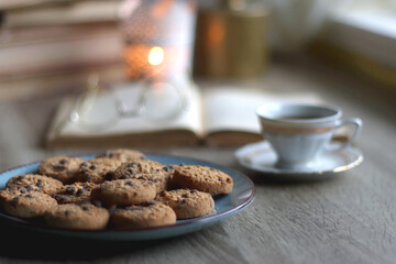 Plate of chocolate chip cookies, stack of vintage books, reading glasses, cup of tea or coffee, lit candle and fairy lights. Hygge at home. Selective focus.