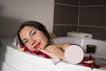 Young smiling brunette woman taking a bath with rose petals at home, doing spa treatments for...