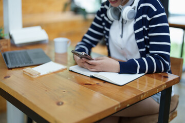 Portrait of a teenage Asian woman using a smartphone, wearing headphones and using a notebook to study online via video conferencing on a wooden desk in library