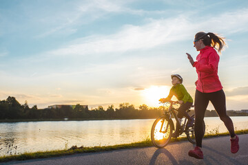 Happy mother and son go in sports outdoors. Boy rides bike in helmets, mom runs on sunny day....