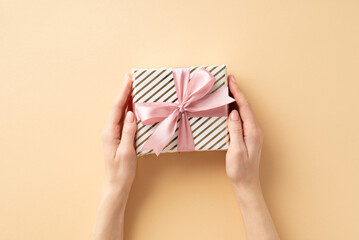 New Year concept. First person top view photo of woman's hands holding stylish giftbox with pink ribbon bow on isolated pastel beige background