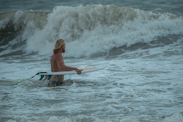 Young handsome man with long hair is on beach with white surfboard in hands