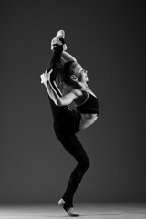 A young flexible girl gymnast in a tracksuit does gymnastic stretching exercises. She threw her leg over her head. Black and white photo on a white background.