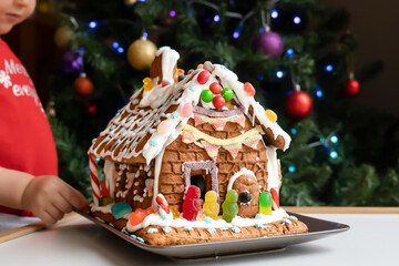 Toddler looking at Homemade gingerbread house decorated with icing, sweets , and jelly candies with blurred Christmas tree on background.