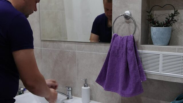 A man in a blue undershirt washes his hands with soap at home in the bath and wipes his hands on a purple towel.