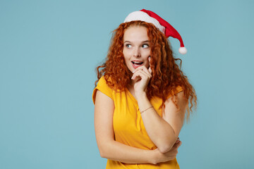 Young smiling fun happy redhead woman 20s wear yellow t-shirt santa claus red hat look aside on workspace area prop up face isolated on plain light pastel blue background. People lifestyle concept.