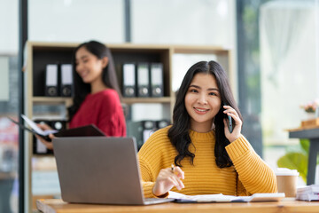 Asian businesswoman chatting on the phone with a customer and looking at a descriptive worksheet on a laptop screen at the office The concept of life in the data analytics office.