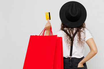Back rear view young woman wear t-shirt hat hold in hand red paper package bags after shopping credit bank card isolated on plain solid white color background studio Black Friday sale buy day concept