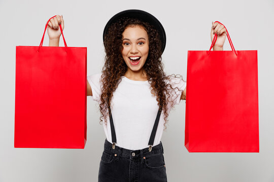 Young Excited Surprised Happy Woman Wear T-shirt Hat Hold In Hand Show Red Paper Package Bags After Shopping Isolated On Plain Solid White Color Background Studio. Black Friday Sale Buy Day Concept.