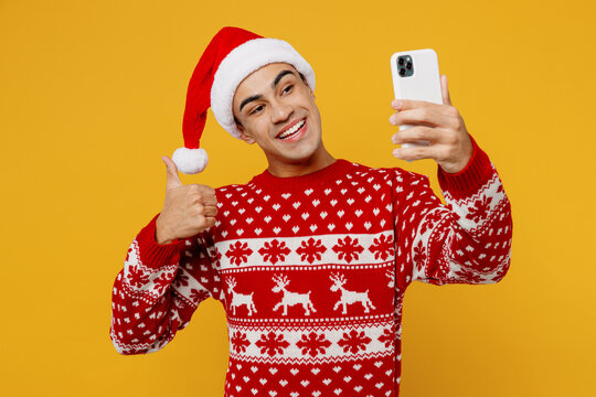 Merry young man wear red christmas sweater Santa hat posing doing selfie shot on mobile cell phone show thumb up isolated on plain yellow background. Happy New Year 2023 celebration holiday concept.