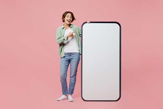 Full body young woman wear green shirt white t-shirt point finger on big huge blank screen mobile cell phone smartphone with workspace copy space mockup isolated on plain pastel light pink background.