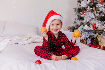 happy little boy in pajamas and Santa hat at home in bed eating tangerines for Christmas