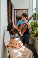 latin young man at home sings and plays guitar to his wife who is pregnant, vertical image