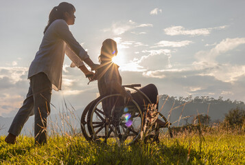Disabled handicapped young woman in wheelchair walking with his care helper in sunset.Silhouette