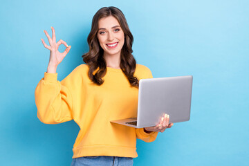 Naklejka premium Photo of young adorable smile cute girl wear yellow sweater hold computer showing okey sign toothy smile recommend app isolated on blue color background