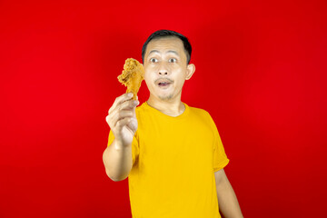 Portrait of young handsome Asian man looking at fried chicken in his hands, wearing yellow shirt and red background.