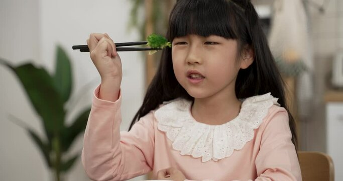 slow motion of upset korean girl complaining with a look of hate while looking at vegetable at dinner table. she picks up the food with chopsticks and puts it down in the bowl