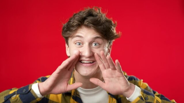 Portrait of young man 20s yelling hey with hands up to mouth isolated on red background. Studio real emotions.