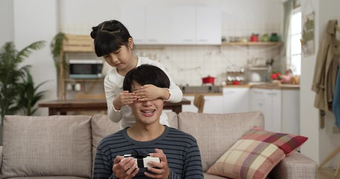 sweet asian girl covering dadâs eyes and hugging him from behind after showing him Fatherâs Day present in the living room at home