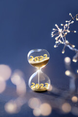 End of the year 2022, Silverster, Happy New Year 2023. Hourglass with numbers 2022 and 2023 and light garland on dark blue background. Hourglass is also known as sandglass, sand glass, sand timer or - obrazy, fototapety, plakaty
