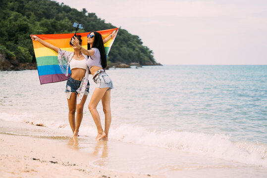 Couple Of Cheerful Women With Rainbow Flag Standing On Beach, Freedom And Love For Same Sex Concept, People With Outdoor Lifestyle, Symbol Of LGBT Community, Summer Vacation And Diversity