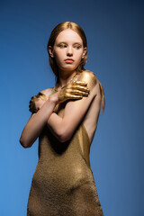Fair haired model in dress and golden paint on hands touching shoulders isolated on blue