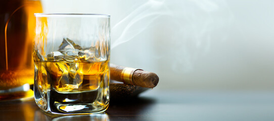 A bottle and a glass of whiskey with ice and a steaming Cuban cigar on a table against a light background. Men's club banner idea. Copy space for text - obrazy, fototapety, plakaty