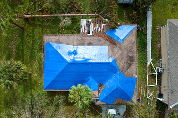 Aerial view of damaged in hurricane Ian house roof covered with blue protective tarp against rain water leaking until replacement of asphalt shingles - obrazy, fototapety, plakaty