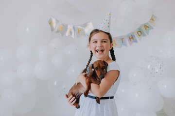 Obraz na płótnie Canvas little girl in a white dress with her beloved dog dachshund in her arms celebrates her birthday