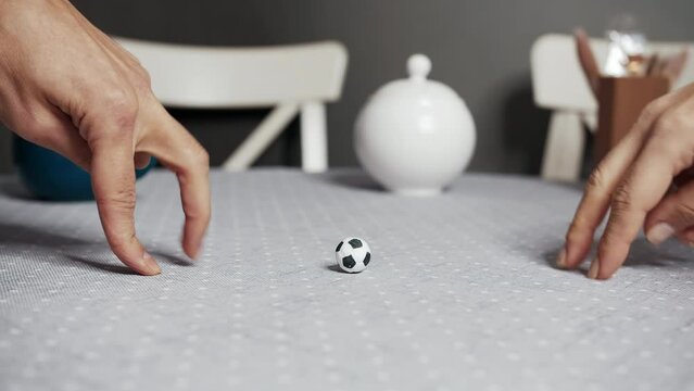 Two people play soccer with their fingers using a small toy ball on the kitchen table.