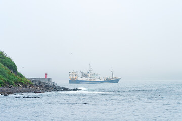 fishing vessel emerges from behind a cape with a lighthouse, sailing into a foggy sea