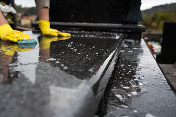 Headstone cleaning on cemetery. Professional in yellow gloves cleans the granite grave, scrubbing with sponge and water. Tombstone preparation for All Saints Day on November 1st