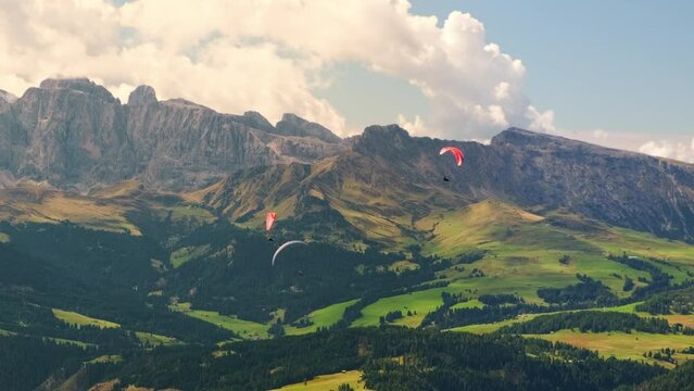 Flying group of paragliders over foresty valley in mountains. Ridge of rocky mountains of Alps under blue sky with clouds aerial view