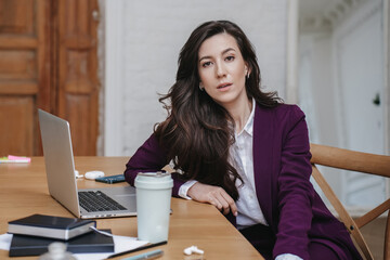 Portrait of attractive businesswoman wis loose wavy hair looking at camera, dressed in burgundy suit, sitting at table with laptop, cup of coffee, notebook, paper sheets. Tired entrepreneur at office - obrazy, fototapety, plakaty