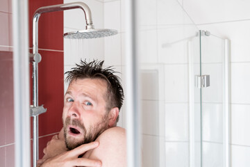Man in the shower under slightly warm water, he freezes and looks miserable. Energy crisis concept.