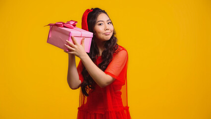 uncertain young asian woman shaking wrapped present isolated on yellow