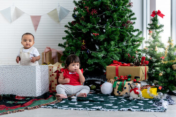 African and Caucasian baby girl, sitting on the floor and eating cookies, with Christmas trees and lots of gift boxes for New Year's Eve celebrations background, to baby and New Year concept.