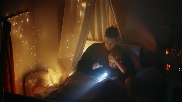 a father lying in bed with his son reads him a fairy tale before going to bed with a lantern. read a book with my son, a lullaby. Father and son in bed. Children's room, to fall asleep, to put to bed