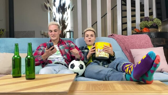 Lucky man celebrating victory, having profit bets at bookmaker website via smartphone app indoors. Soccer fans watching live broadcast sport match on TV, cheering favorite football team at home