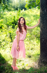 Naklejka na ściany i meble A girl in a fabulous image with elf ears in a pink dress, in a sunny forest and in backlight. Selective focus. Blurred background.