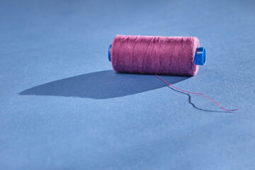 An oblong spool of sewing thread of a beautiful purple color lies with unwound thread on a harmonious blue background in bright sunlight with contrasting shadows. Needlework, sewing