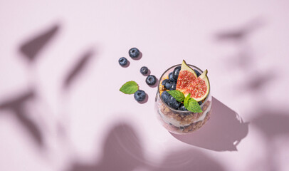 Delicious natural yogurt with granola, figs and blueberries in a glass on a pink background with shadows.