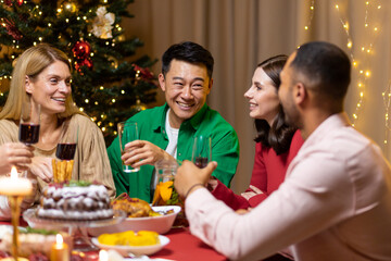 Happy young interracial people celebrating new year at home at festive table near Christmas tree. They hold glasses in their hands, laugh, communicate, rejoice