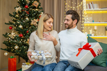 Obraz na płótnie Canvas Family couple in love man and woman on Christmas celebrating New Year holidays and exchanging gifts sitting on sofa at home near Christmas tree in living room.