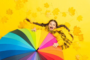 A child holds onto an autumn multicolored umbrella lying on a yellow background with maple fallen leaves. A little girl in a raincoat with a scarf is enjoying the autumn weather.
