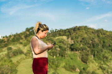 beautiful young latina woman, on top of a mountain with her cell phone in her hand, interacting on social networks. in the background a big hill and a blue sky.