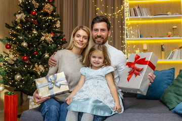 Obraz na płótnie Canvas Portrait of mature family with daughter for Christmas, man woman and child looking at camera with gifts and smiling near Christmas tree at home sitting on sofa in living room.