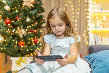 A little girl is watching a video on a smartphone for Christmas, her daughter is celebrating the New Year online near the Christmas tree