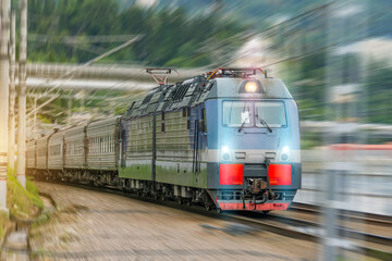 Powerful electric locomotive brightly turned on headlights pulls a train of passenger cars, with motion blur effect.