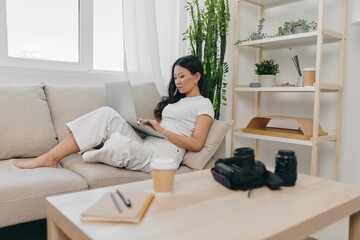 Asian woman photographer lying at home on the couch and reviewing photos from the camera in laptop for color correction retouching, work as a freelance business photographer for yourself home office